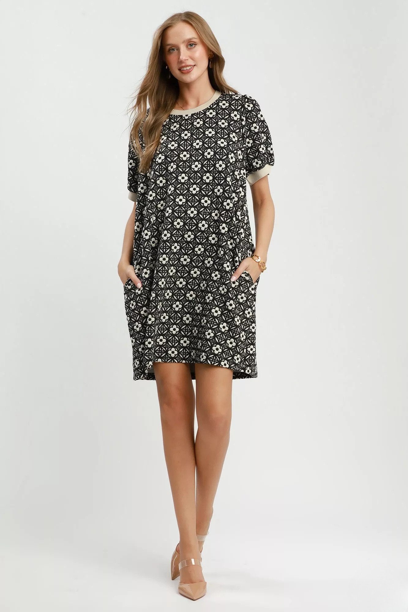 Model wearing a black floral knit swing dress with short puff sleeves, contrast trim, side pockets, and a relaxed silhouette styled with nude heels..