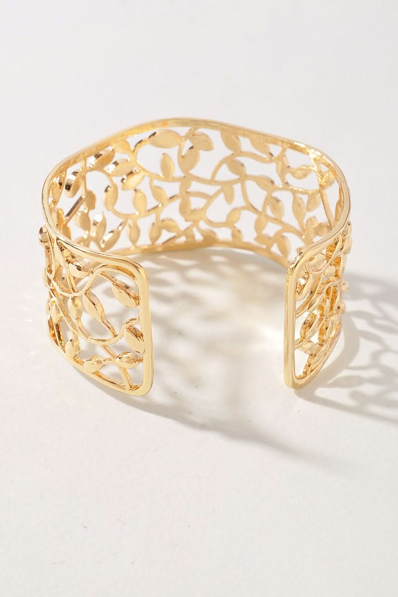A gold-toned open cuff bracelet with an intricate leaf filigree pattern, displayed on a clean white surface with soft shadows.
