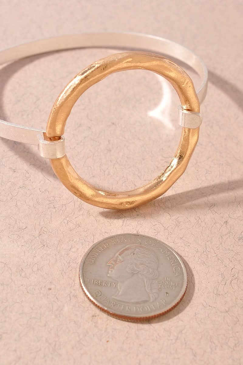 A gold-toned circular bracelet bangle with a white inner band, shown on a textured beige surface next to a US quarter coin for scale.