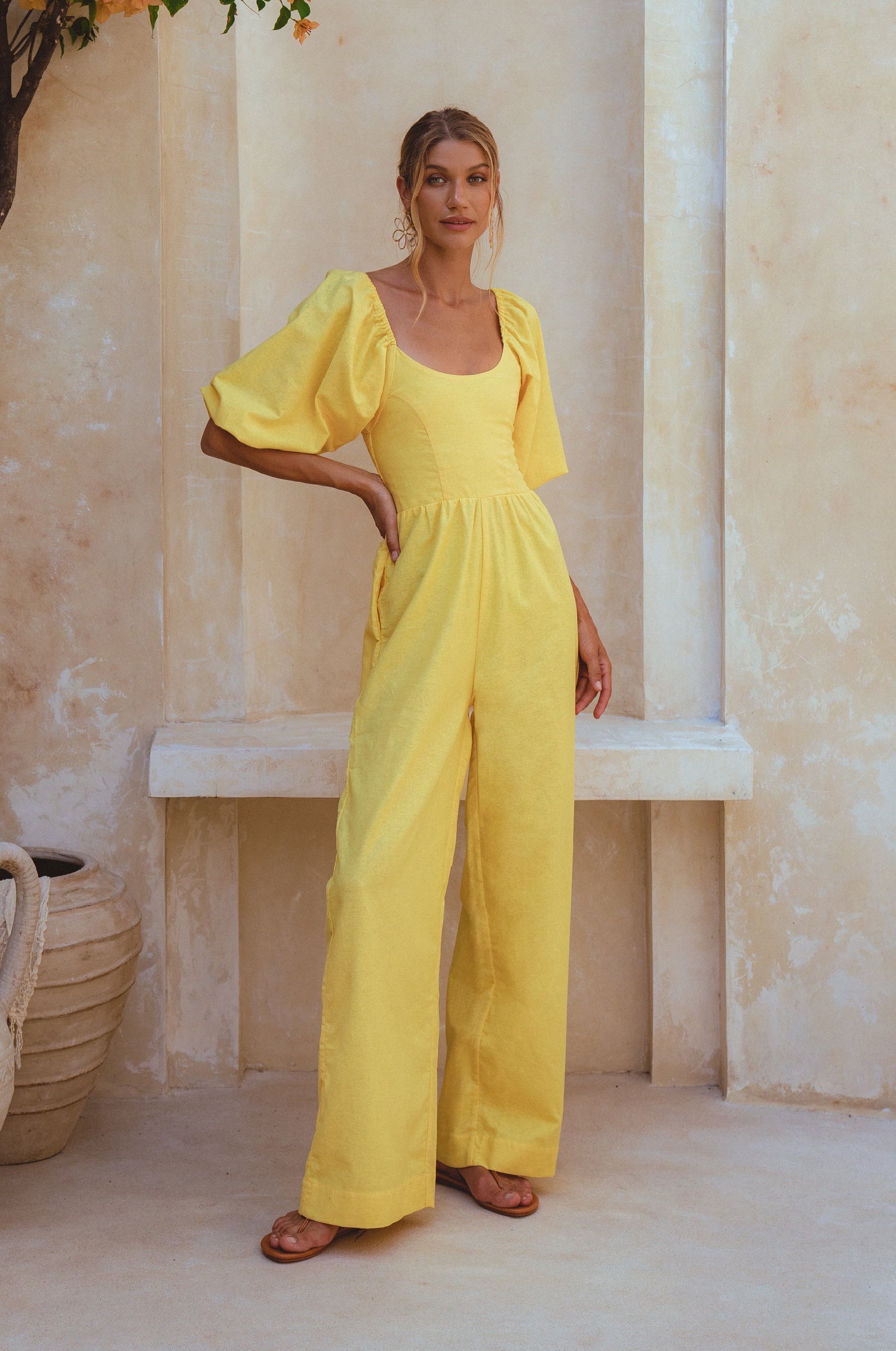 Woman wearing a yellow linen jumpsuit with puff sleeves and round neck standing against a neutral wall, front view