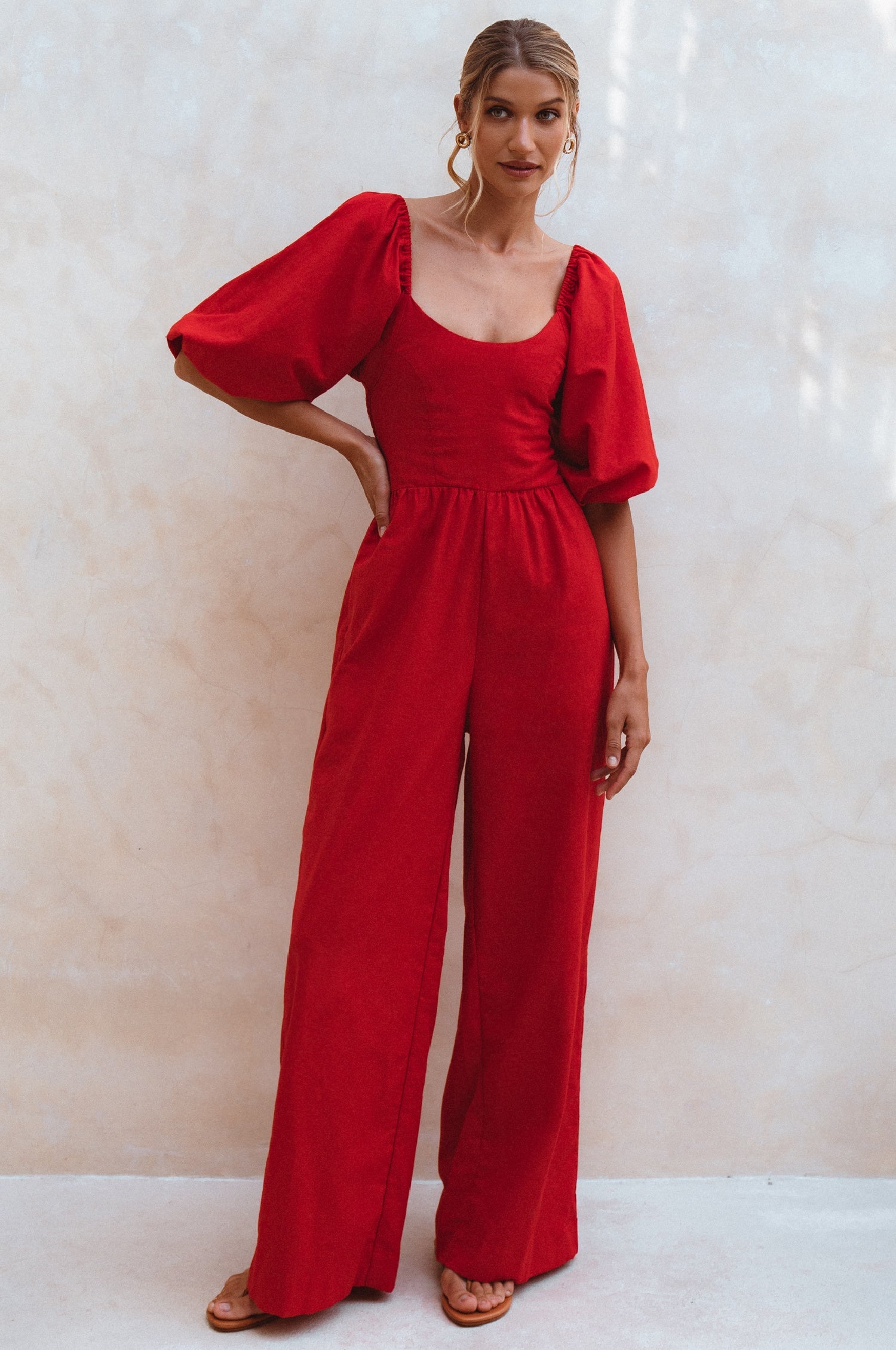 Woman wearing a red linen jumpsuit with puff sleeves and round neck standing against a plain wall, front view