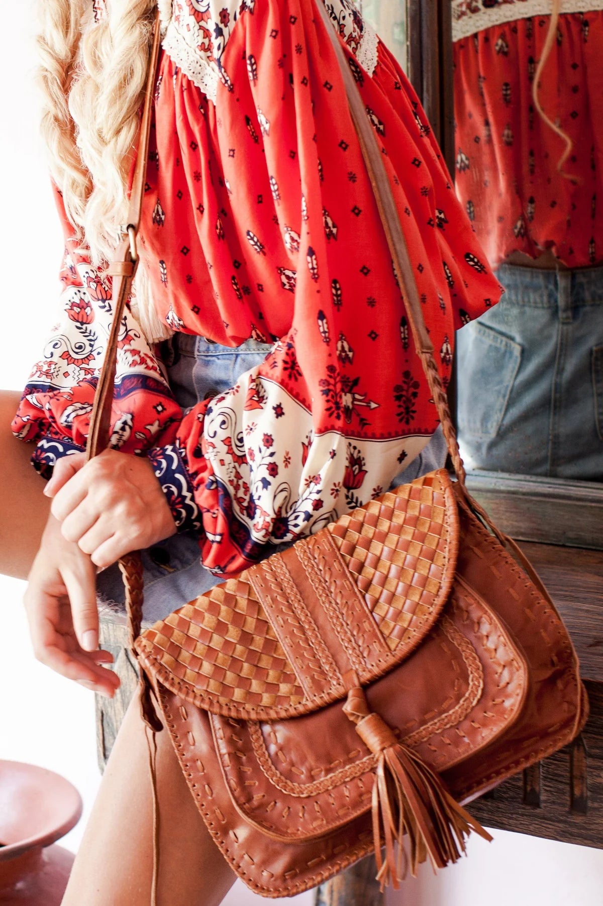 A close-up lifestyle shot of a person wearing a red patterned bohemian blouse and holding a brown woven leather crossbody bag with a tassel.