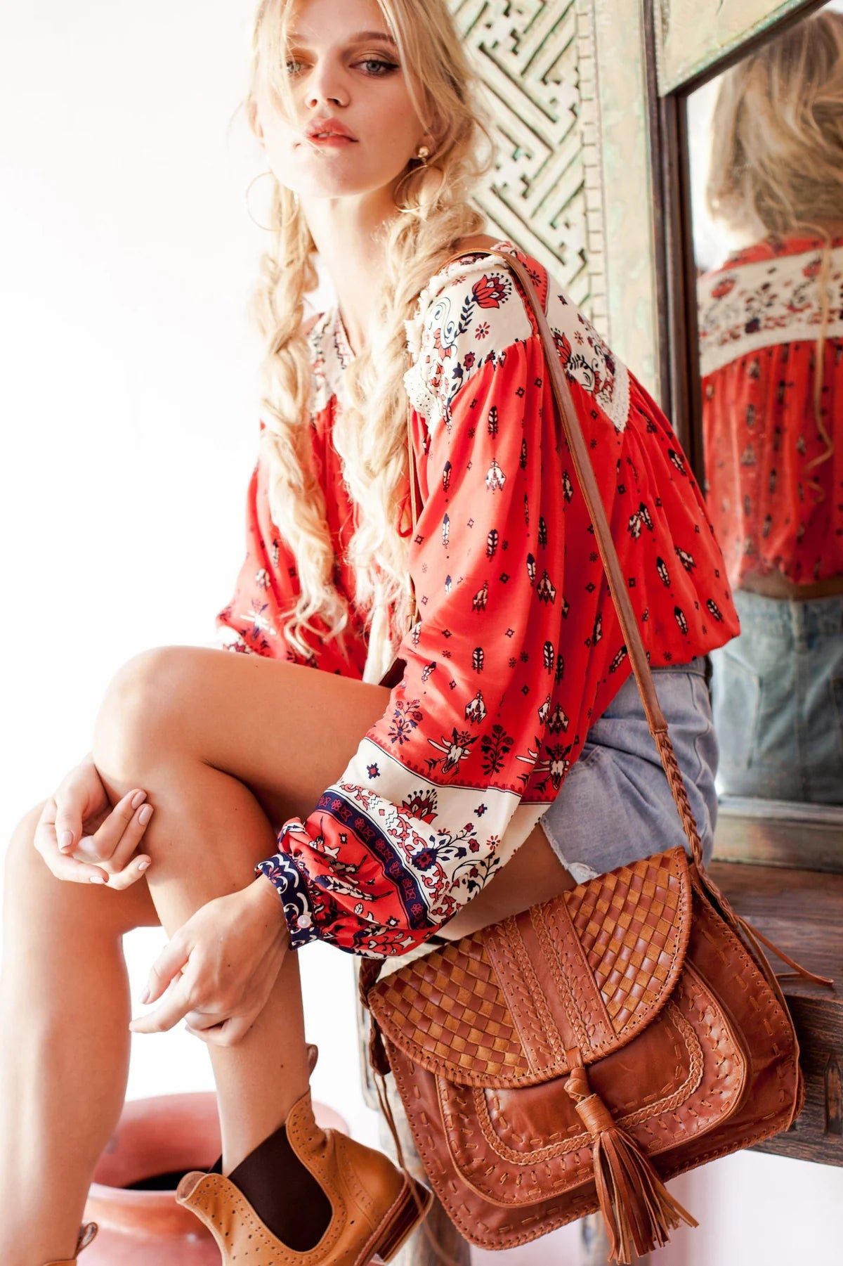 Woman in a red and white patterned blouse with a brown leather bag with tassel sitting on a chair.