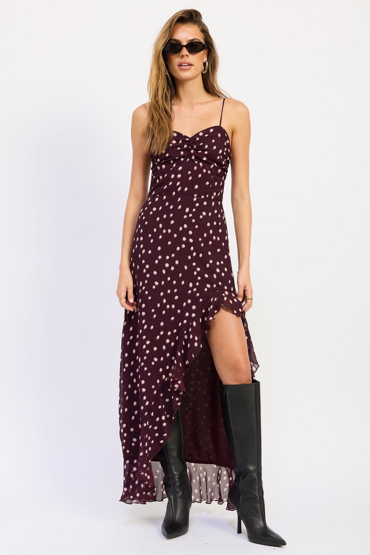 Woman wearing polka dot maxi dress with spaghetti straps and ruffled asymmetric hem in Wine against white background.
