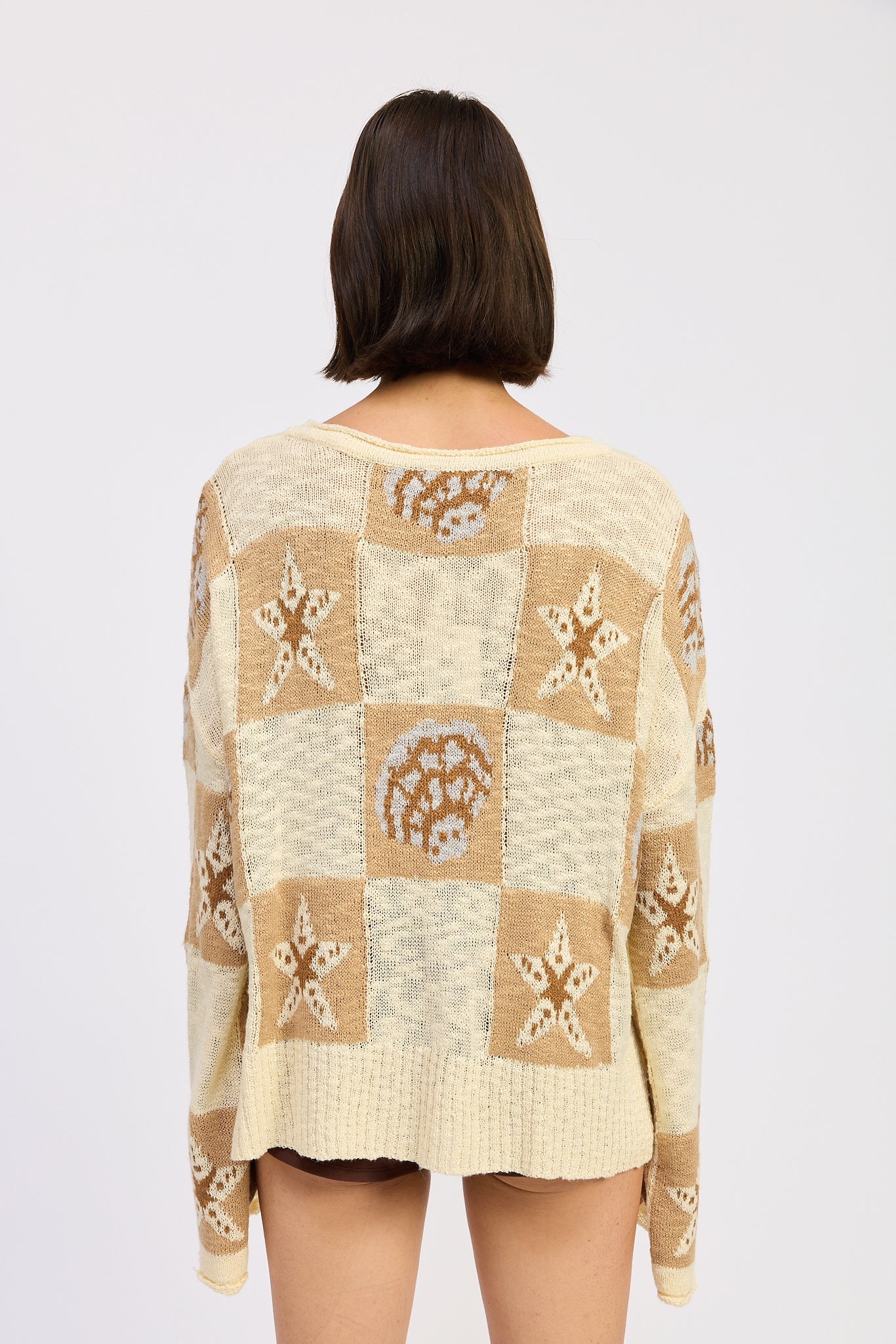 Woman wearing a cream and taupe patchwork knit sweater with star and seashell motifs, backview. and brown knee-high boots on a white background