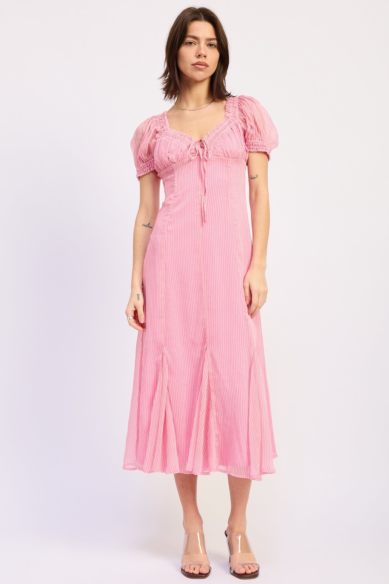 Model wearing a pink pinstripe midi dress with puff sleeves, a tie front neckline, fitted bodice, and flared skirt styled with clear wedge sandals.