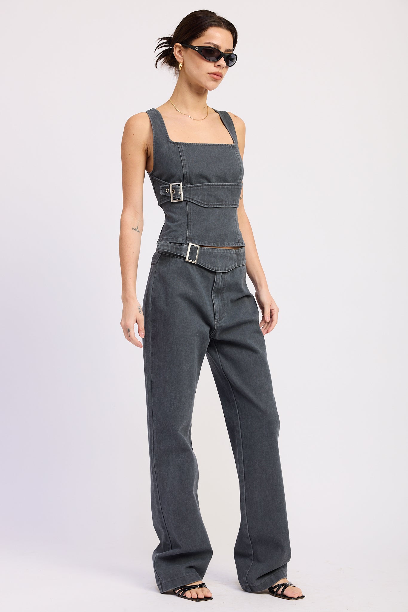 Woman wearing a washed charcoal denim corset top with buckle detail front view with sunglasses on a plain background front angle view
