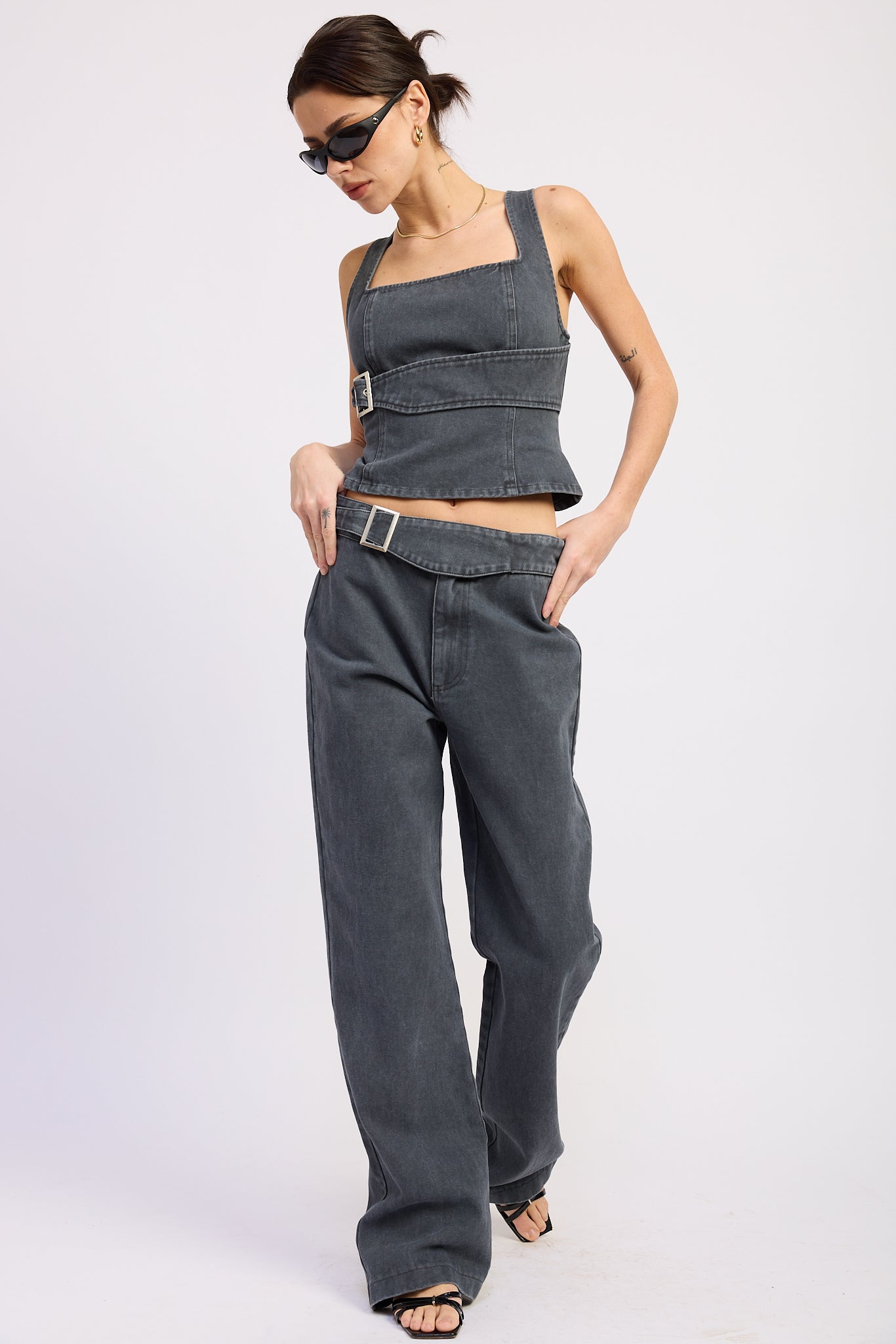 Woman wearing a washed charcoal denim corset top with buckle detail front view with sunglasses on a plain background