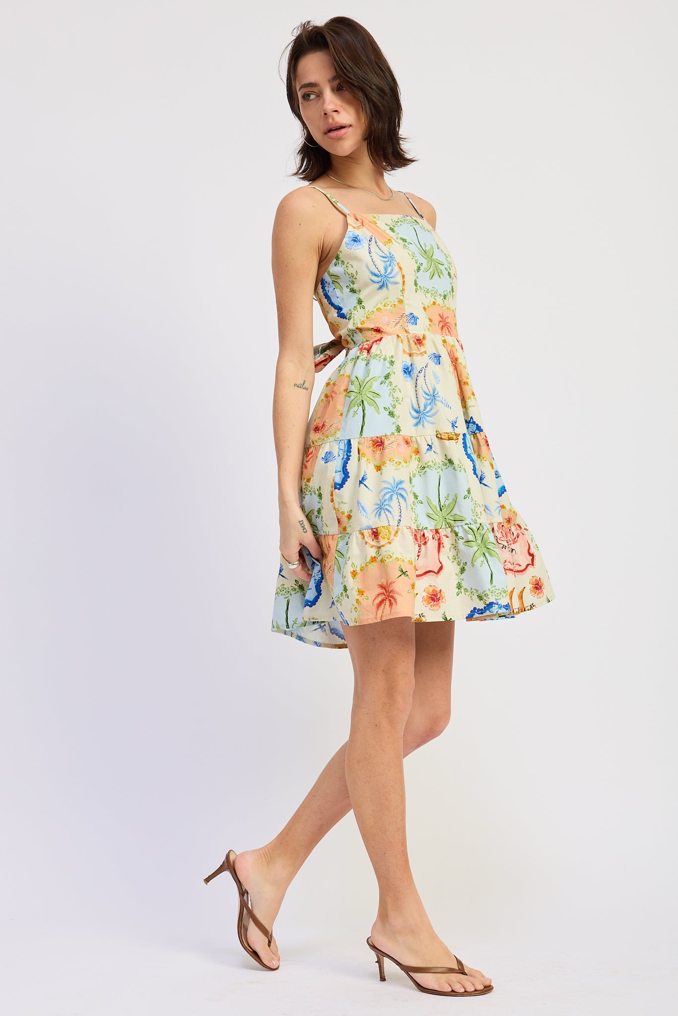 Woman wearing a scarf-inspired print blend of palm motifs and delicate detailing for a vibrant colorful floral tiered mini dress on a white background, side view