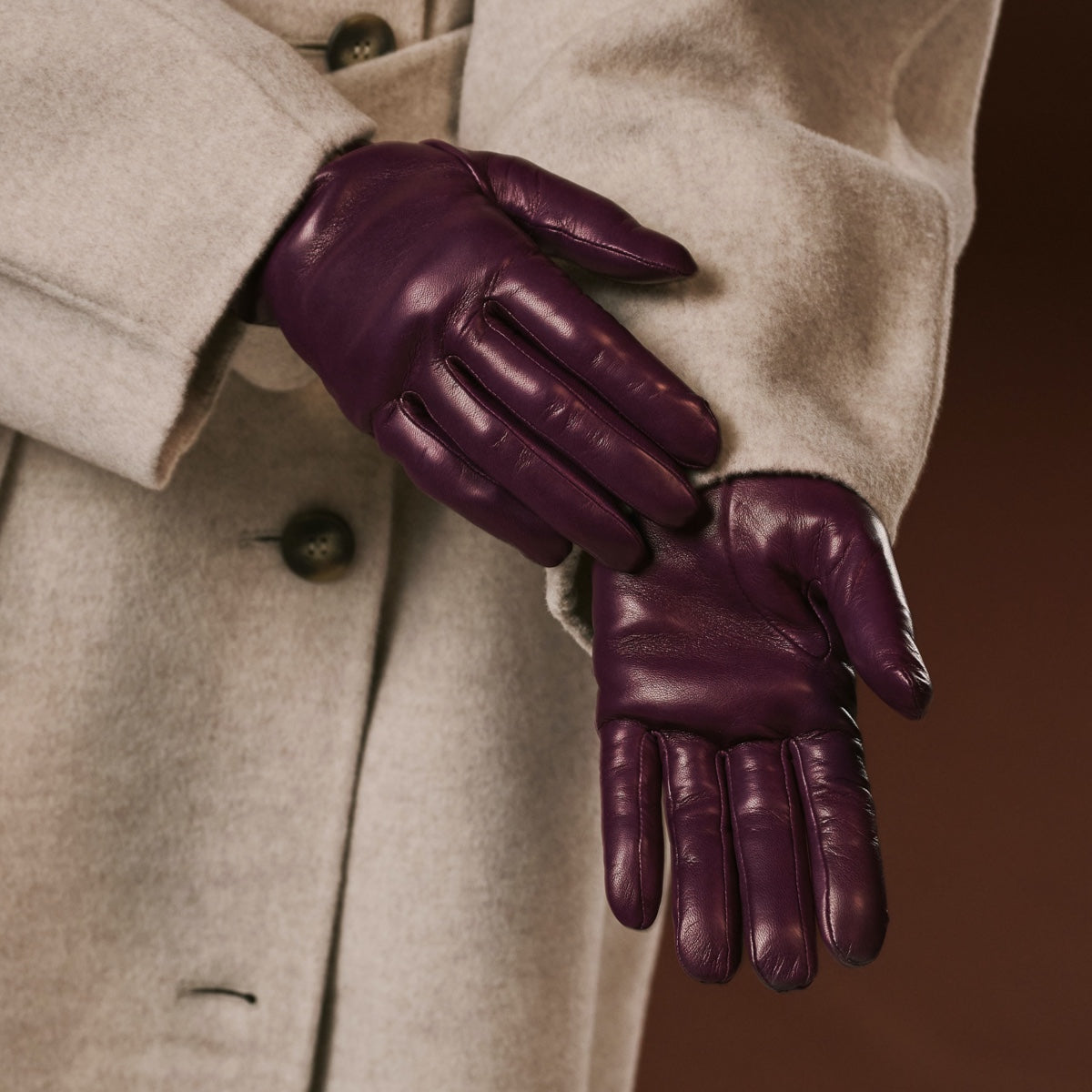 Purple leather gloves worn with a beige coat against a brown background