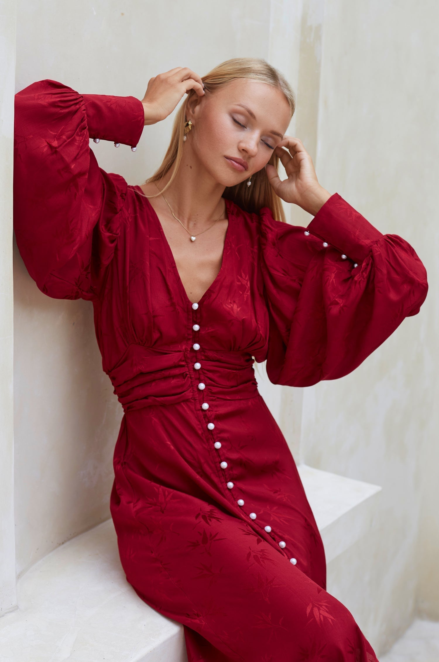 Woman in a red Woman midi dress with pearl buttons, V-neckline, balloon sleeve, and fitted detailing on the front sitting on a white bench.