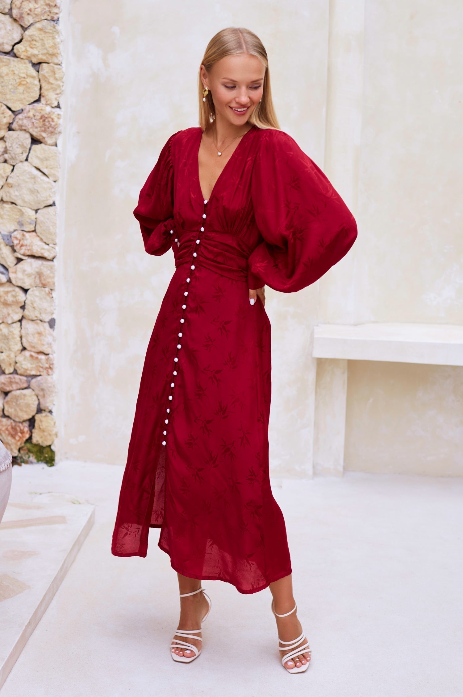 Woman wearing a red midi dress with pearl buttons, V-neckline, balloon sleeve, and fitted detailing on the front in a white room, 3/4 view