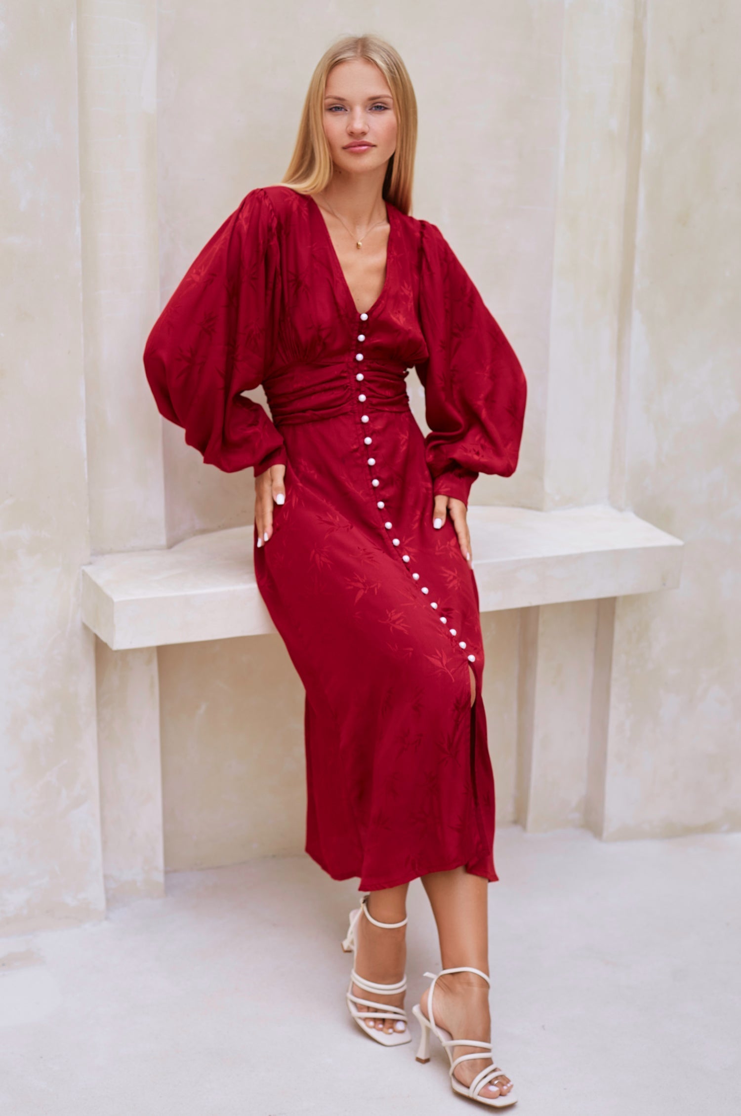 Woman wearing a red midi dress with pearl buttons, V-neckline, balloon sleeve, and fitted detailing on the front leaning against a bench in a minimalistic setting, front view