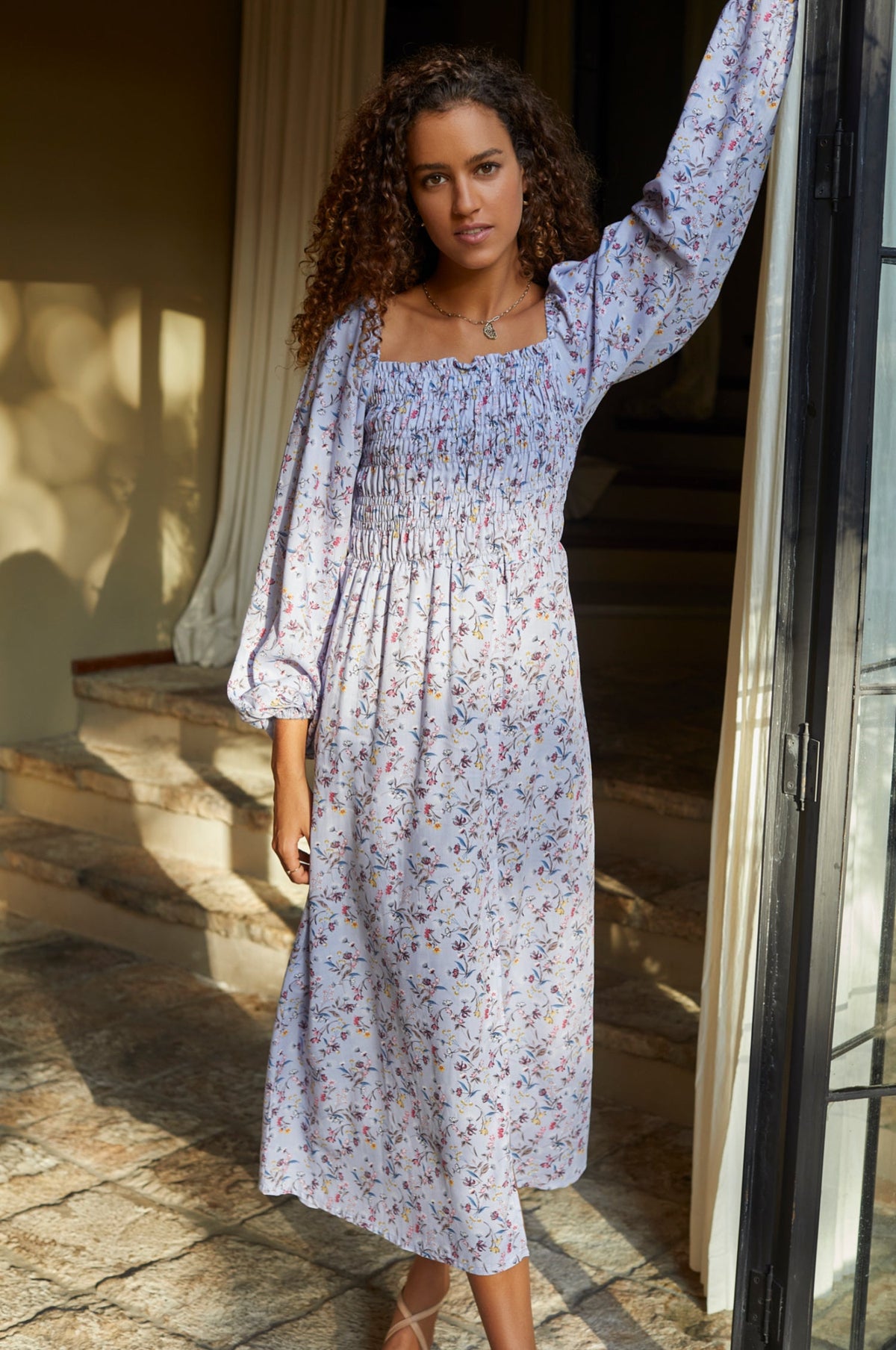Woman in a lavender floral midi dress standing on a stone path with a building in the background