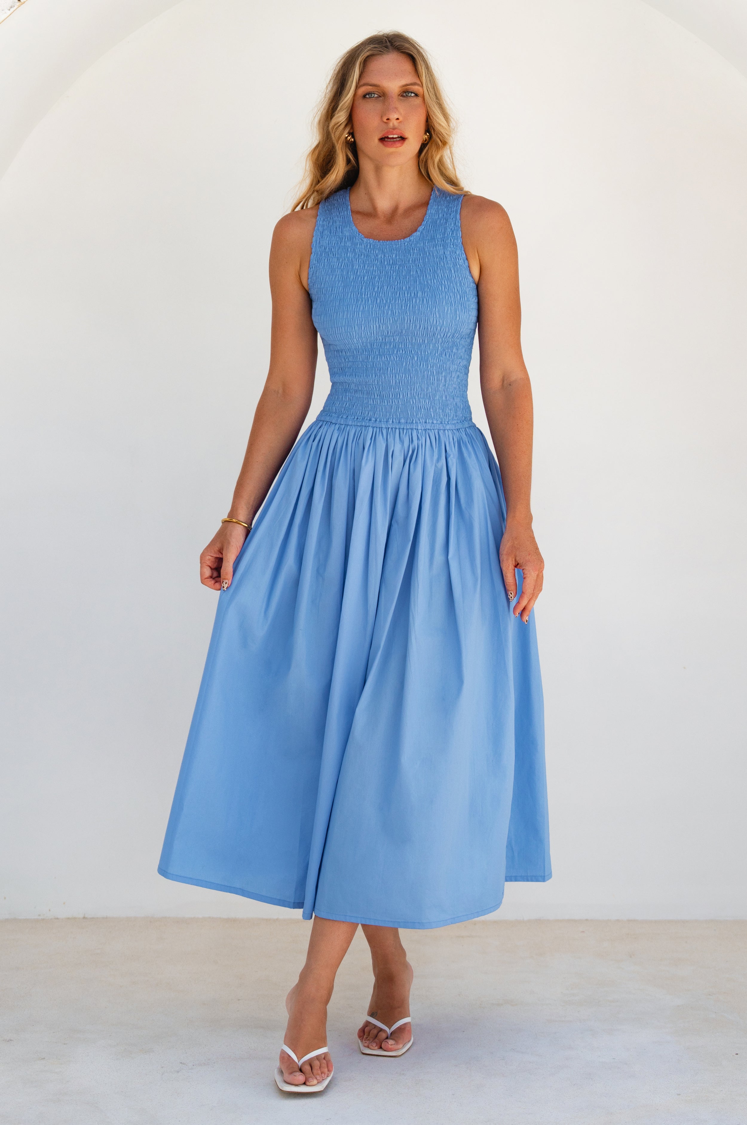 Woman wearing a light blue cotton sleeveless midi with scoop neck, shirred bodice and flare skirt against a white background, front view.