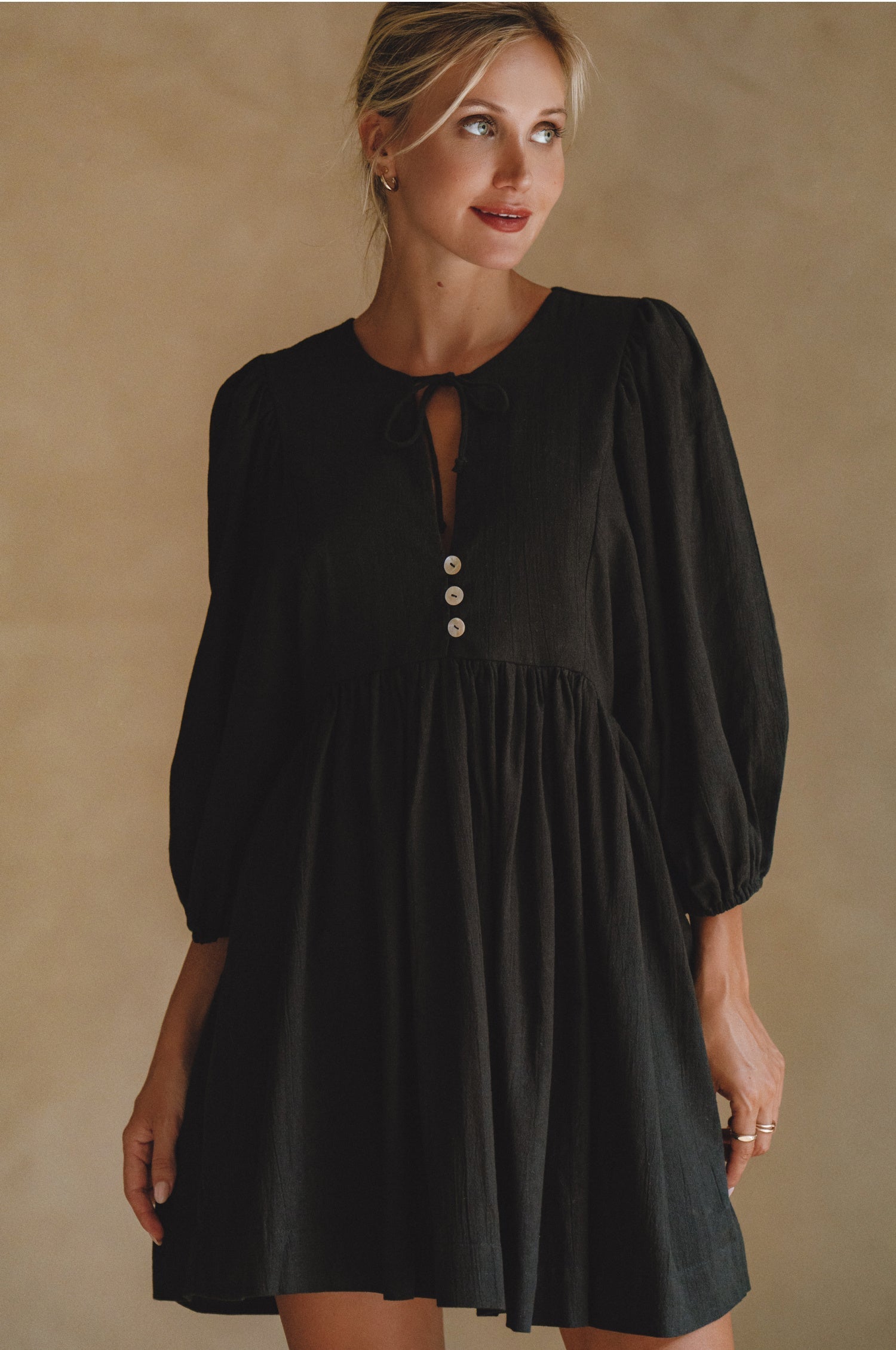 Woman wearing black colored linen mini dress or lightweight crinkle linen in a long-sleeve babydoll silhouette against a neutral background, front view