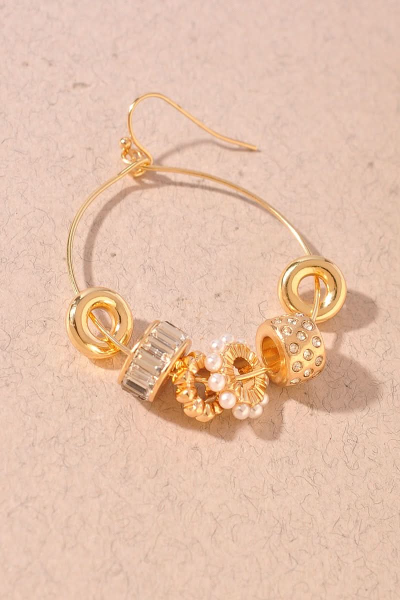 Gold hoop earring with decorative charms, including a textured gold bead, a clear crystal bead, and a pearl bead, displayed on a textured beige background.