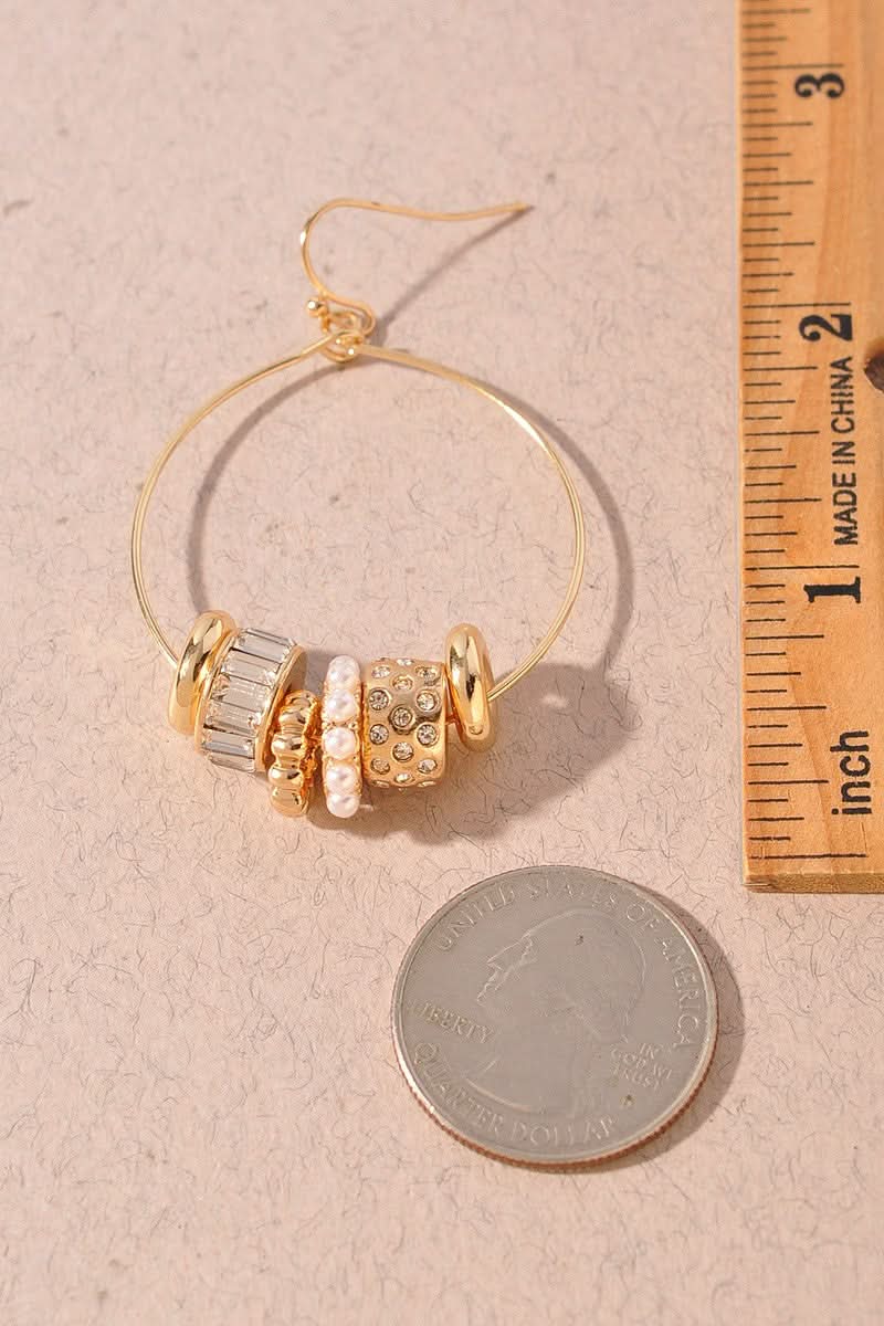 Gold hoop earring with three decorative bead charms, shown next to a US quarter for scale and a wooden ruler on a textured beige background.