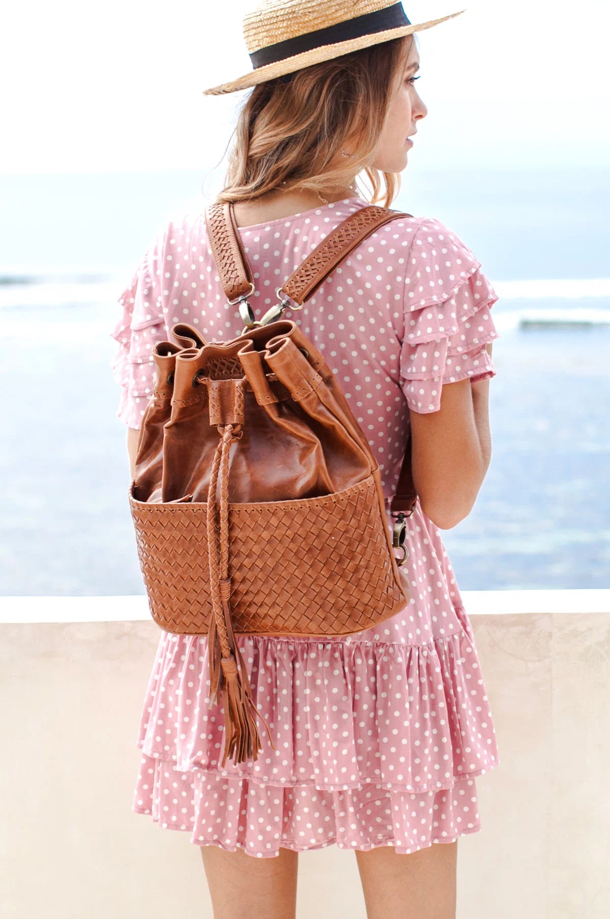 A woman wearing a pink and white polka dot dress and straw hat, seen from behind, carrying a camel woven leather bucket backpack with a tassel drawstring and braided straps against a beach background.