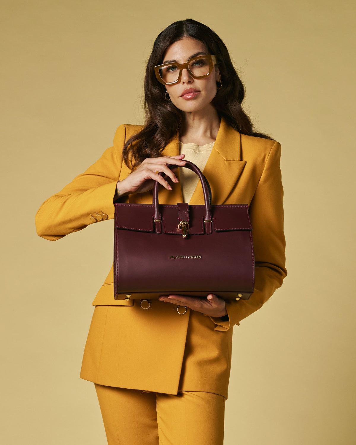 Woman in a mustard suit holding a burgundy handbag against a beige background