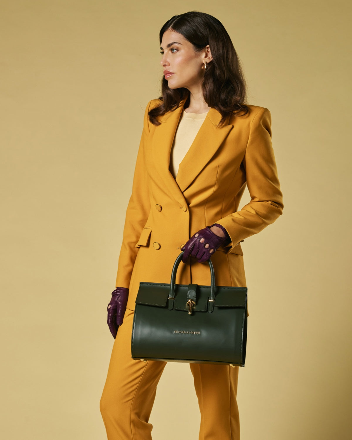 Woman in mustard yellow suit holding a green leather handbag against a beige background