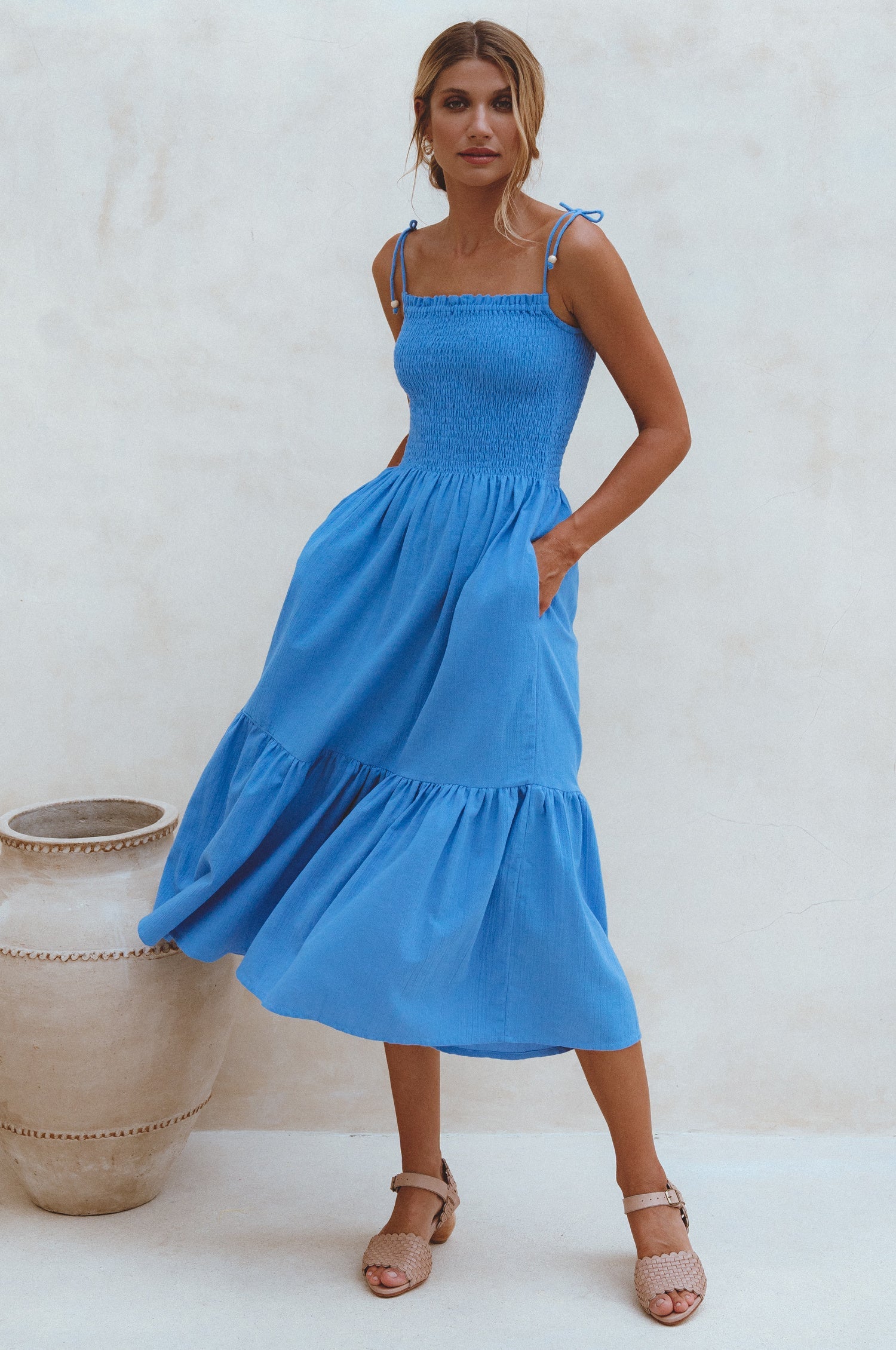 Woman wearing a blue linen tiered midi dress with wooden bead adorned spaghetti straps and smocked bodice with her hands in the pockets standing against a white wall, front view