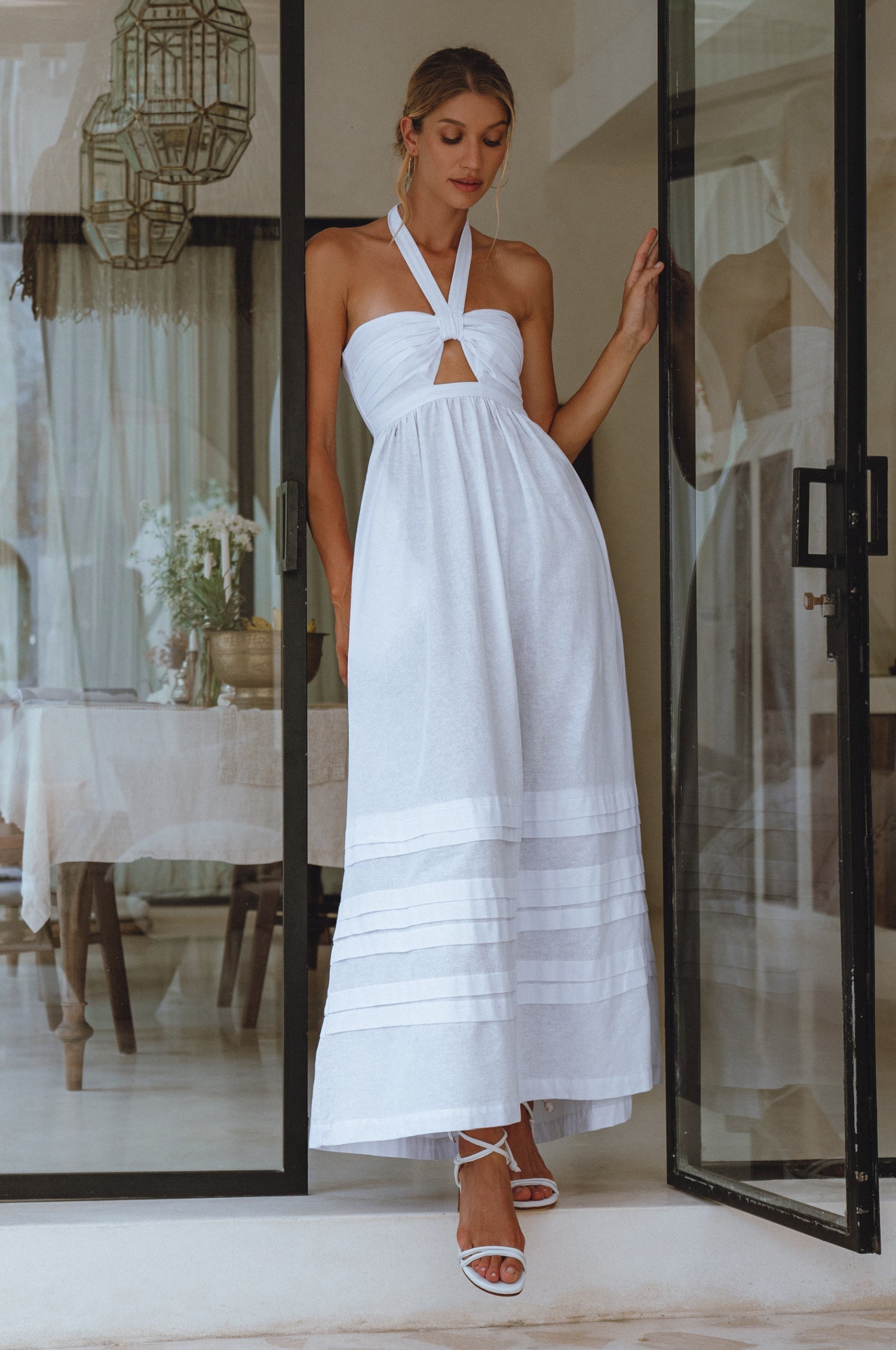 Woman wearing a white linen maxi dress with halter neck, front cut out and beautiful fold details at hem standing in a modern interior setting