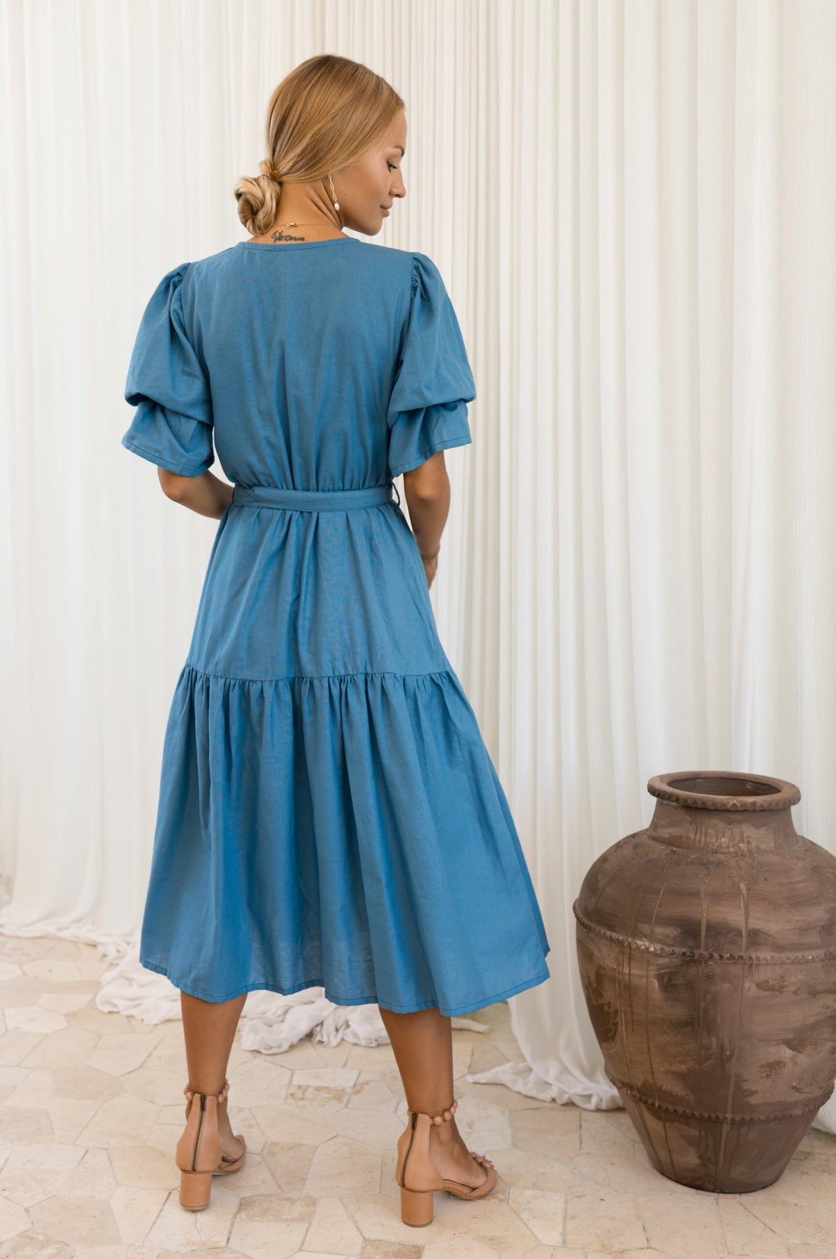 Woman wearing a blue dress standing next to a large vase against a white curtain background back view