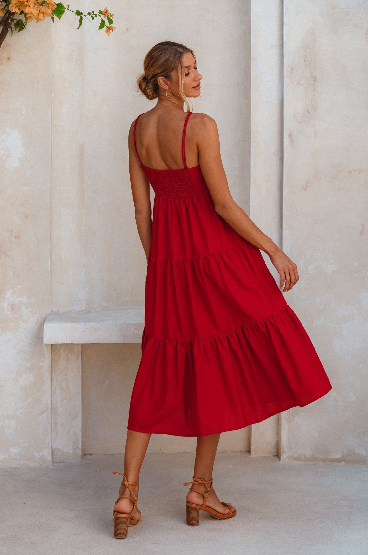 Model wearing a red linen tiered midi dress with dual adjustable spaghetti straps standing against a light-colored wall, back view