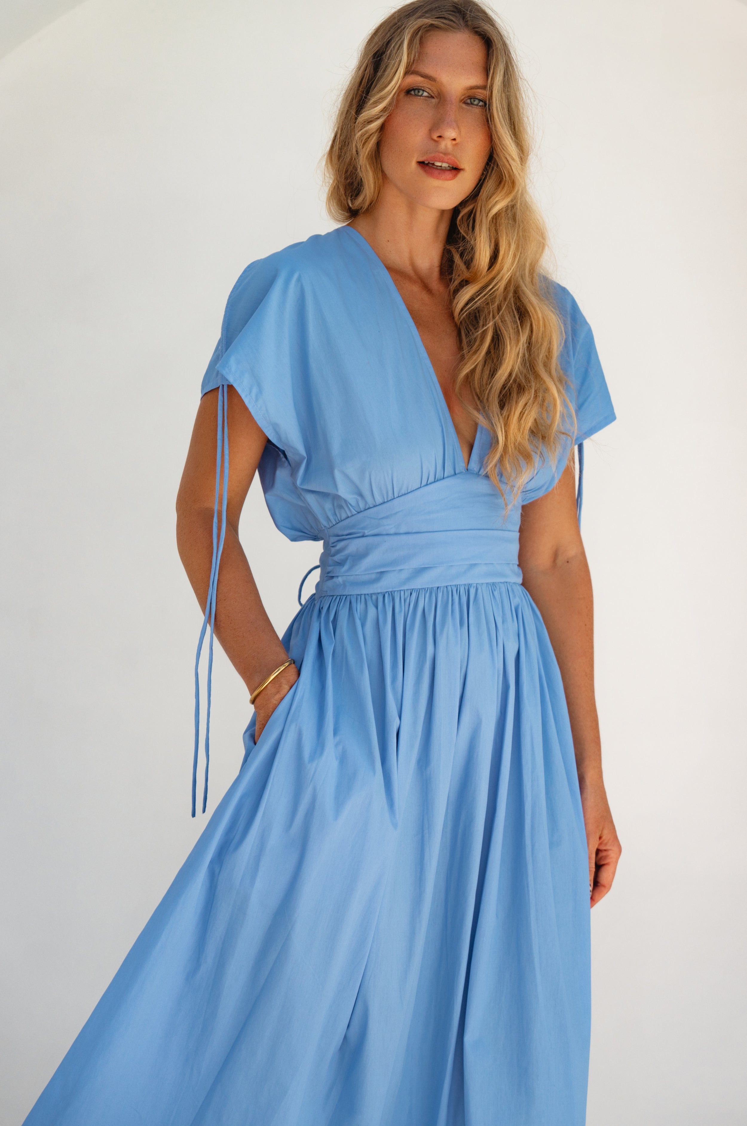 Woman wearing a blue maxi dress with sleeves un-gathered, deep v-neckline and hand in pocket against a white background