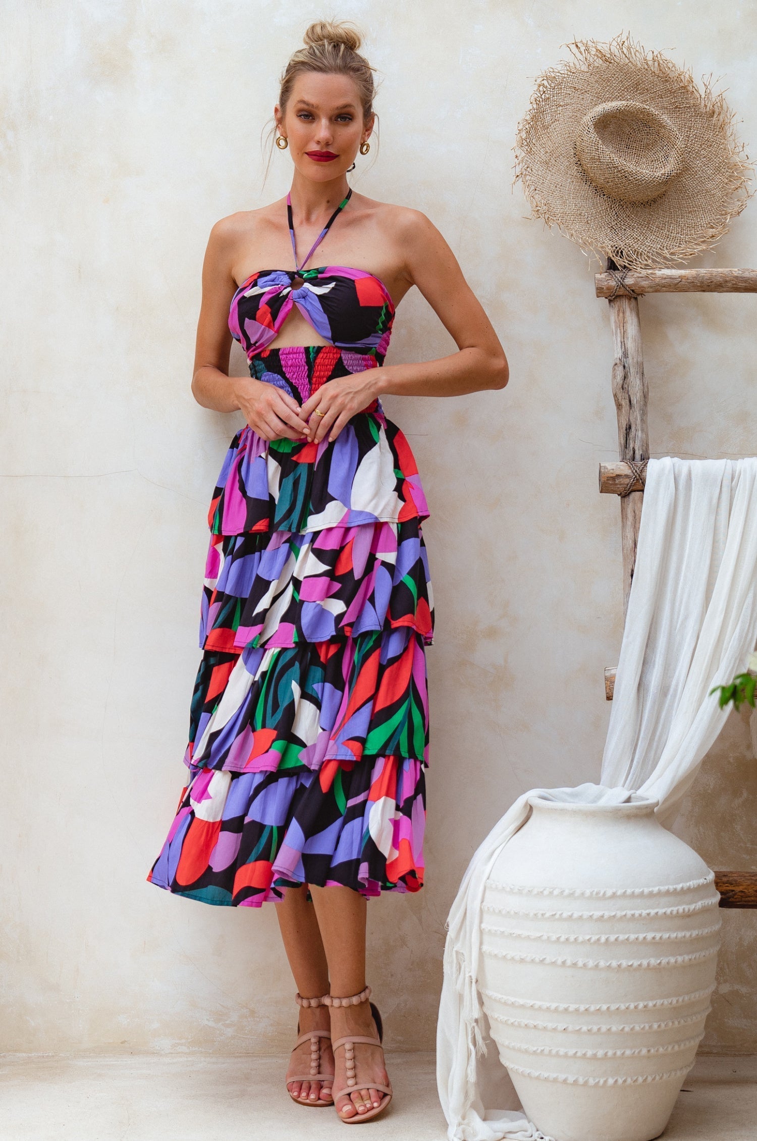 Woman wearing a colorful abstract floral haltered midi dress standing next to a white vase and straw hat on a wooden stand.
