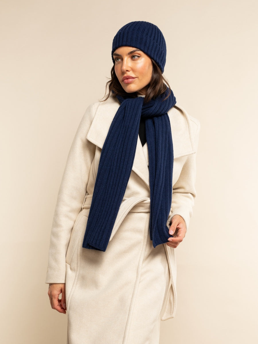Woman wearing a white coat, navy blue cashmere beanie, and scarf against a beige background
