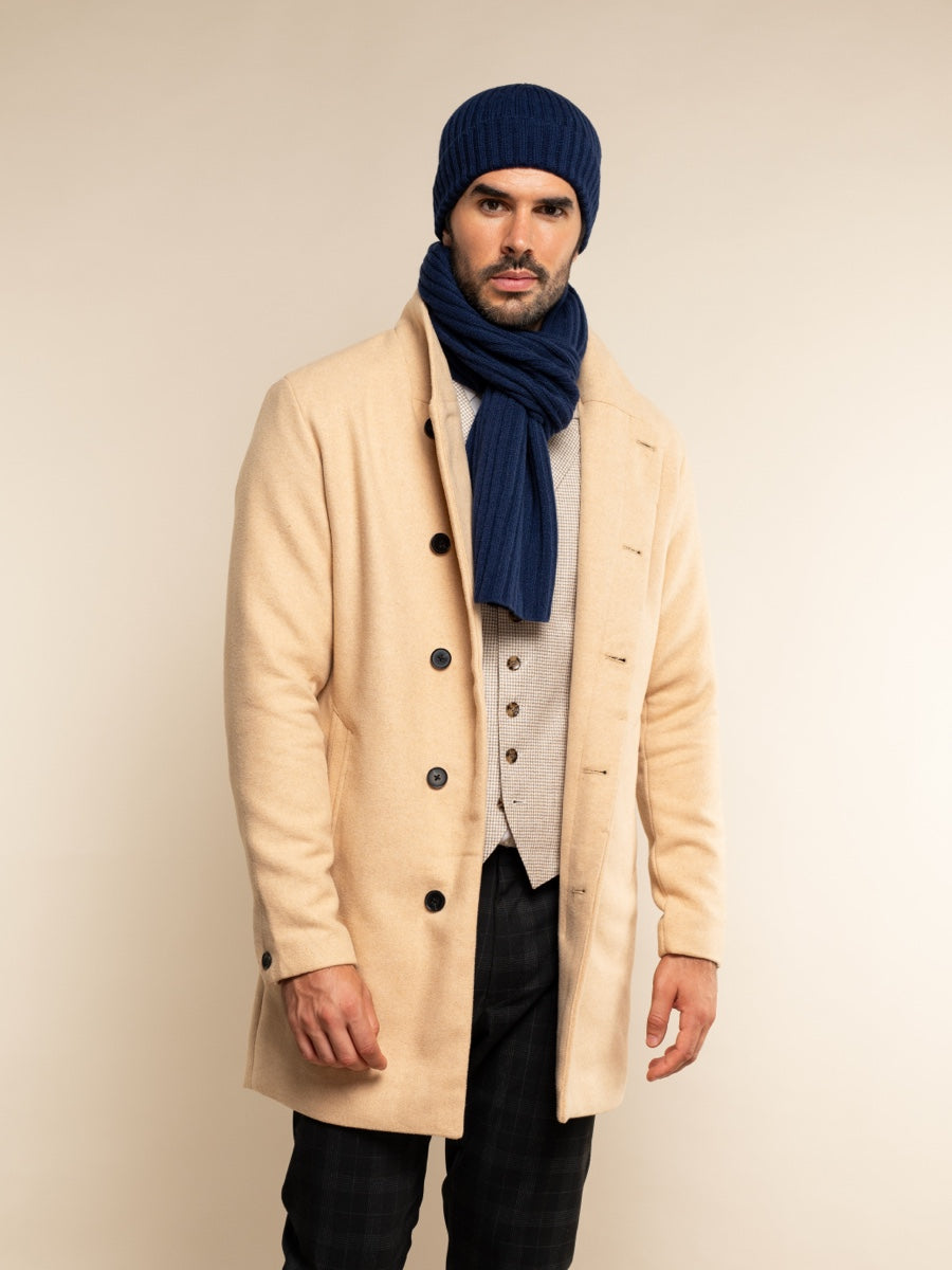 Man wearing a beige coat, navy beanie, and scarf against a beige background