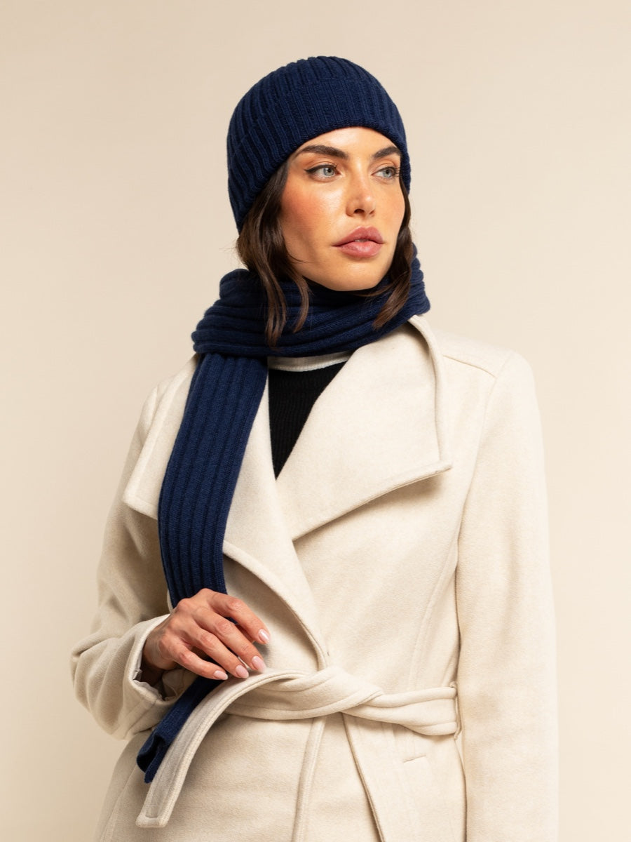 Woman wearing a navy blue cashmere knit beanie and scarf with a beige coat against a plain background