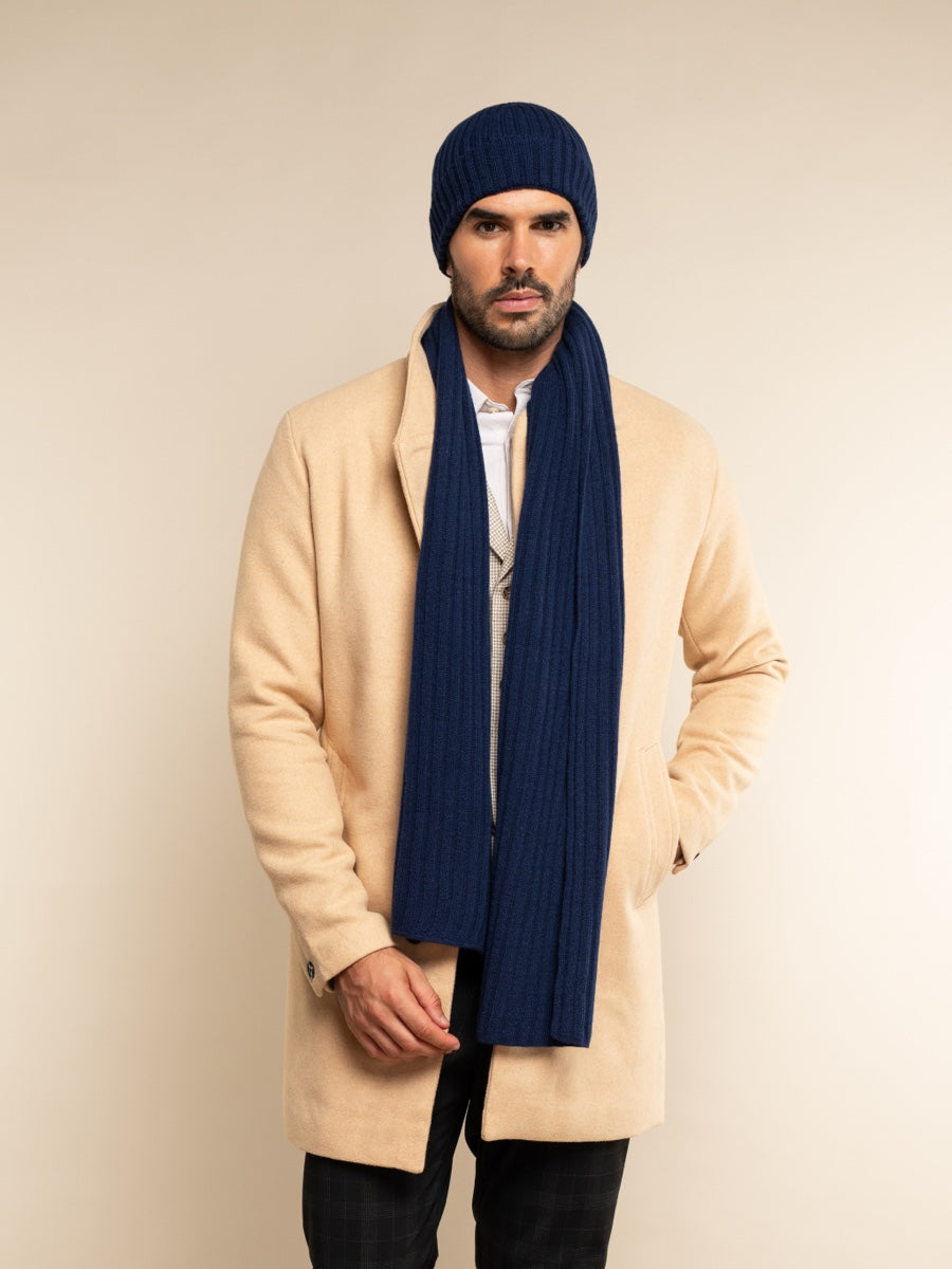 Man wearing a beige coat, cashmere navy beanie, and scarf against a beige background