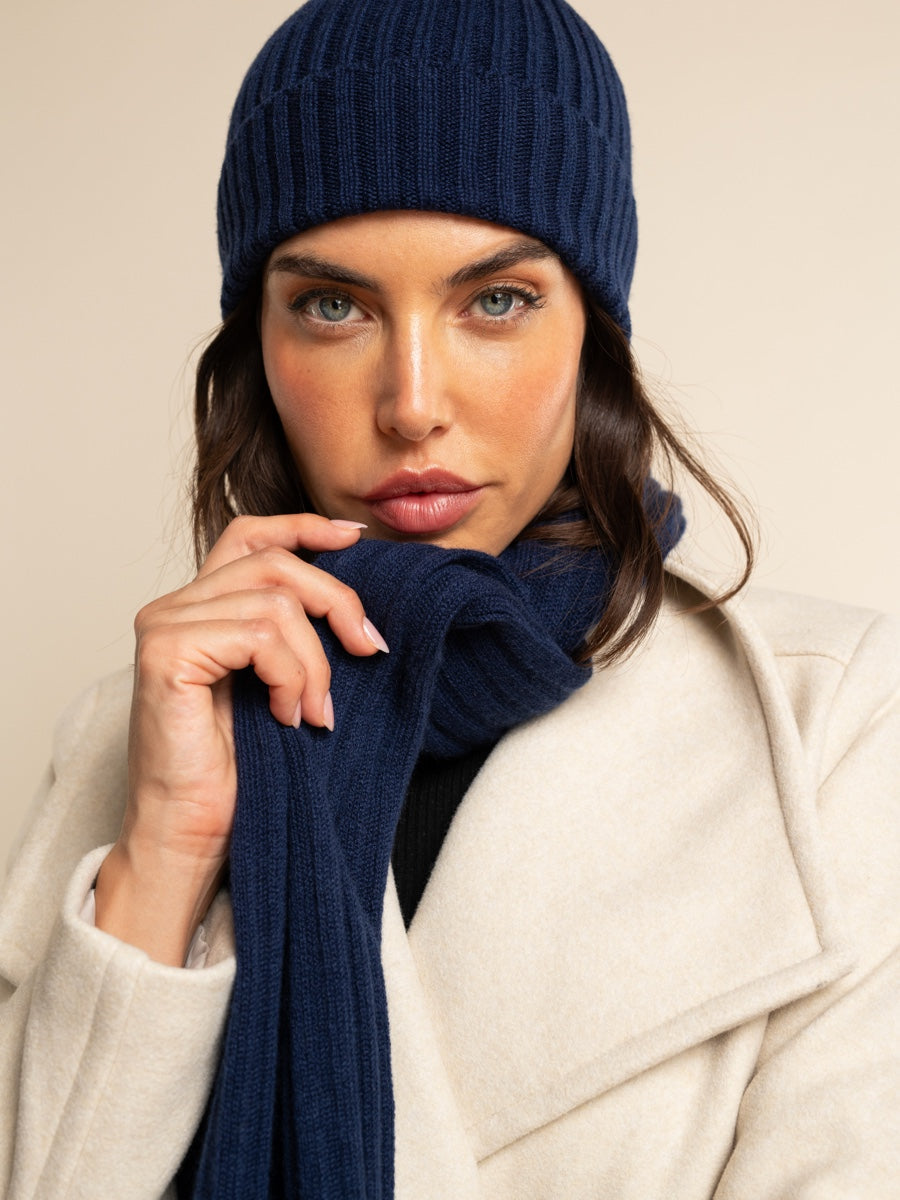 Woman wearing a navy blue knit cashmere hat and scarf against a beige background