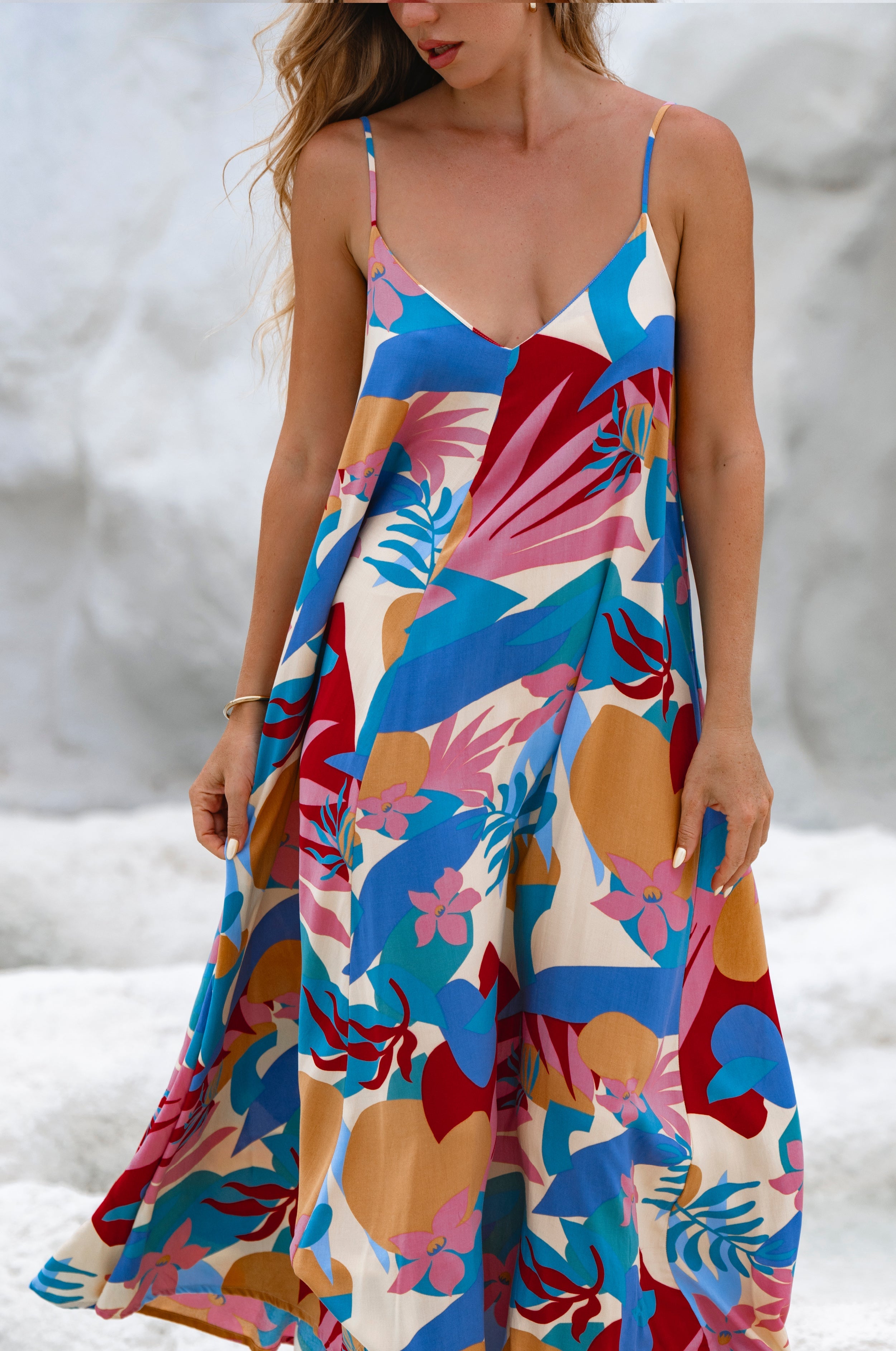 Woman wearing a blue colorful abstract floral flowy maxi dress with deep v-neckline and spaghetti straps with a blurred background