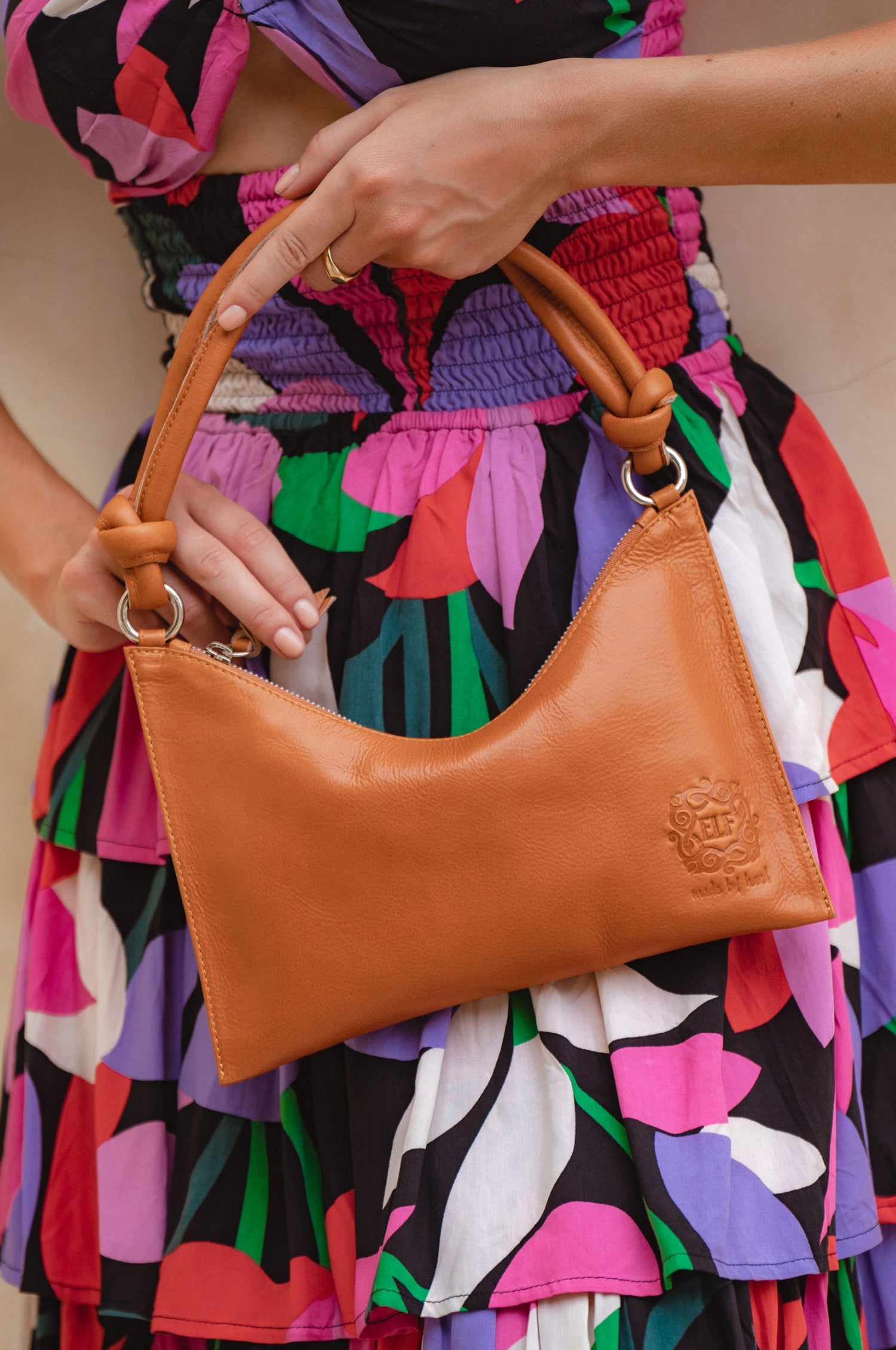 Person holding a tan leather handbag with a colorful dress in the background