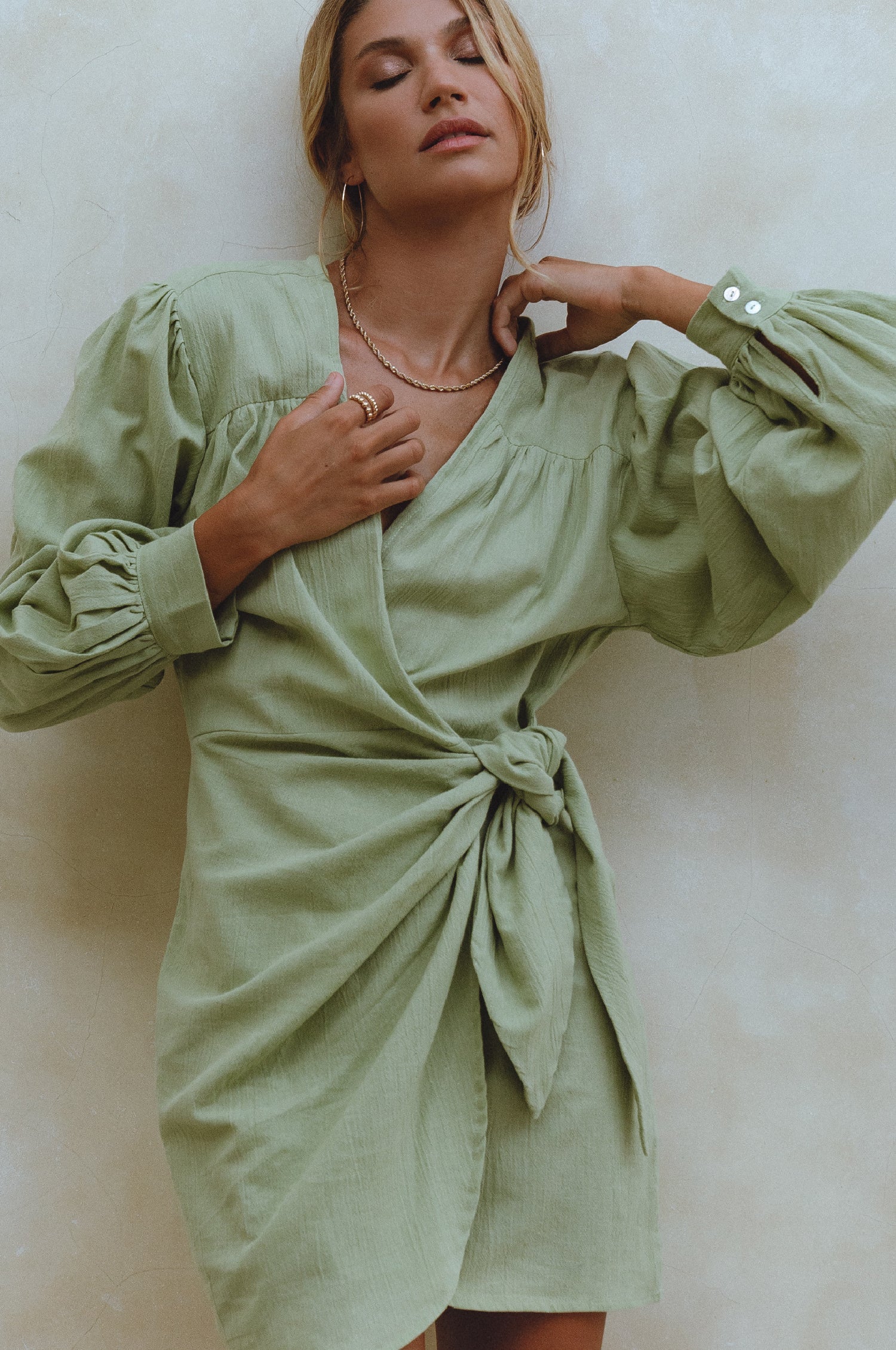 Woman wearing a sherbert green crinkle linen mini dress with adjustable wrap, a V-neckline and billowy sleeves, against a white background, front view