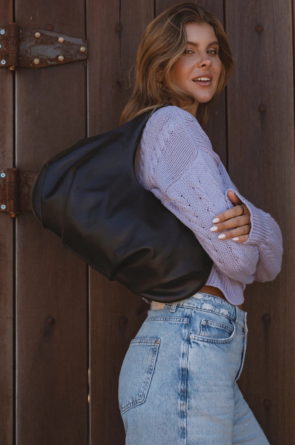 Woman wearing a large black leather slouchy bag and light purple sweater against a wooden door.
