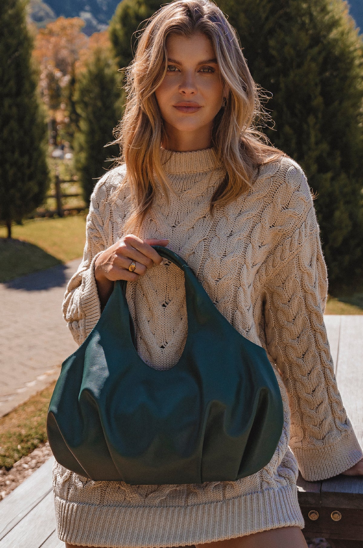 Woman holding a green leather bag outdoors with trees and a path in the background