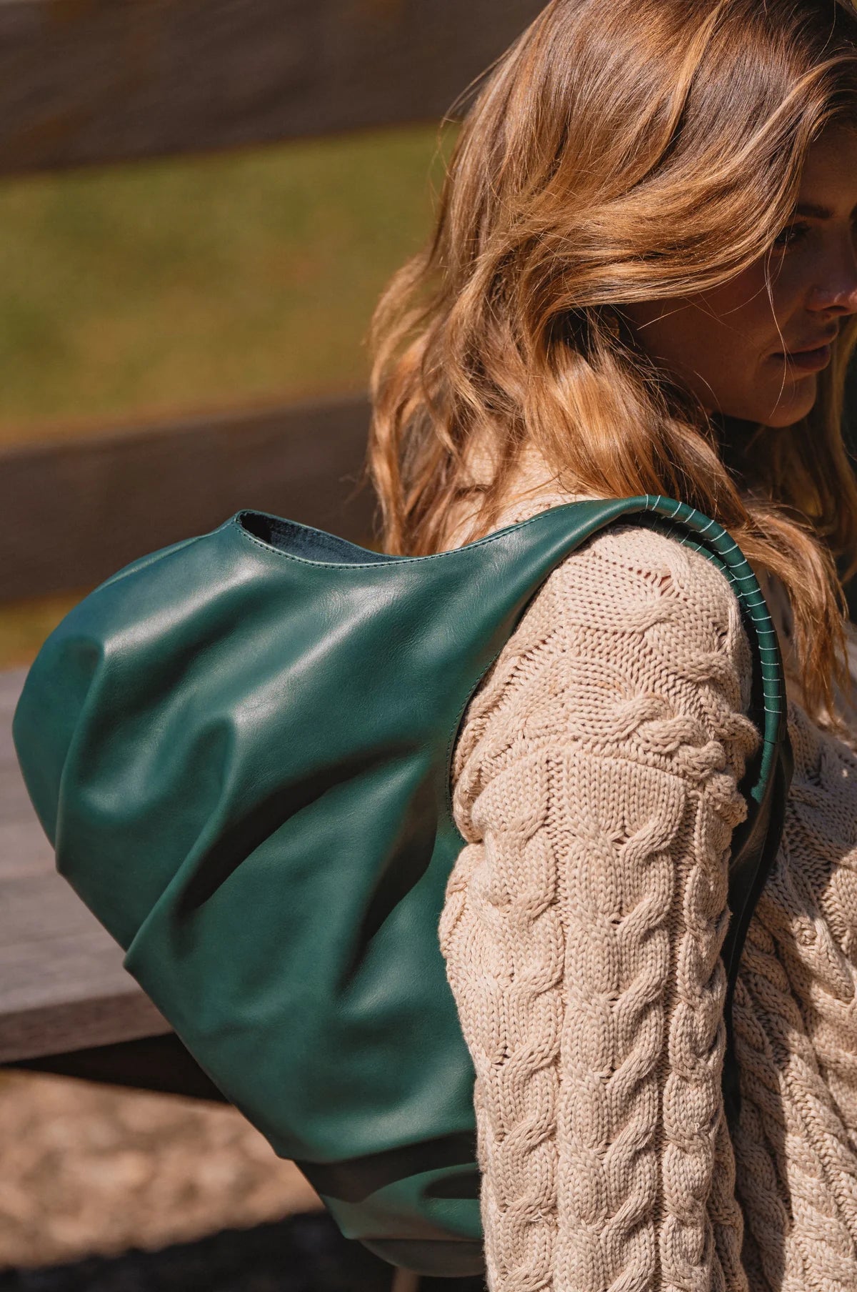 Person wearing a beige knitted sweater with a green leather bag over their shoulder outdoors.