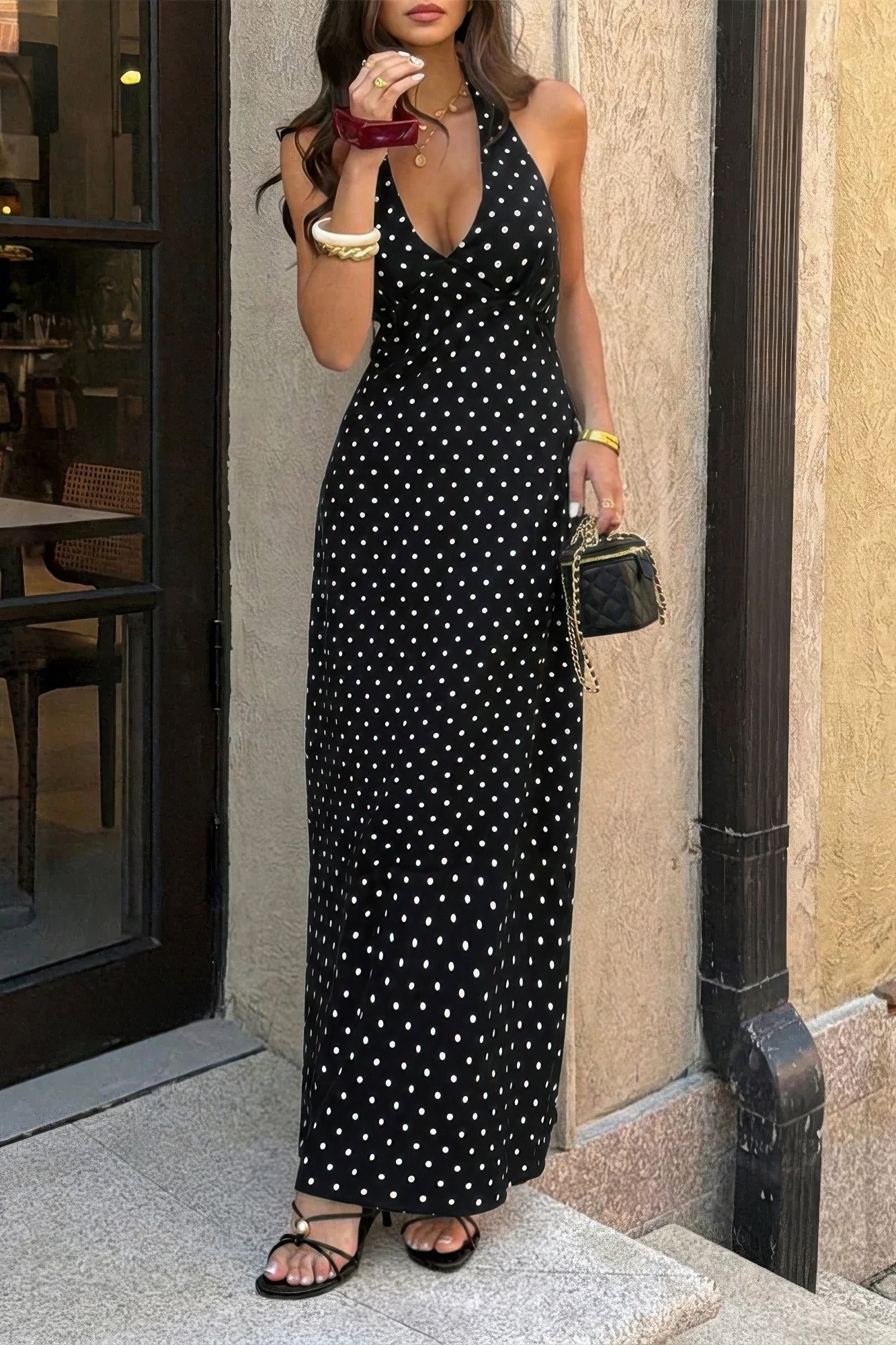 Woman wearing long black with maxi dress with white polka dots with halter and deep v-neckline, outdoors in front of restaurant window