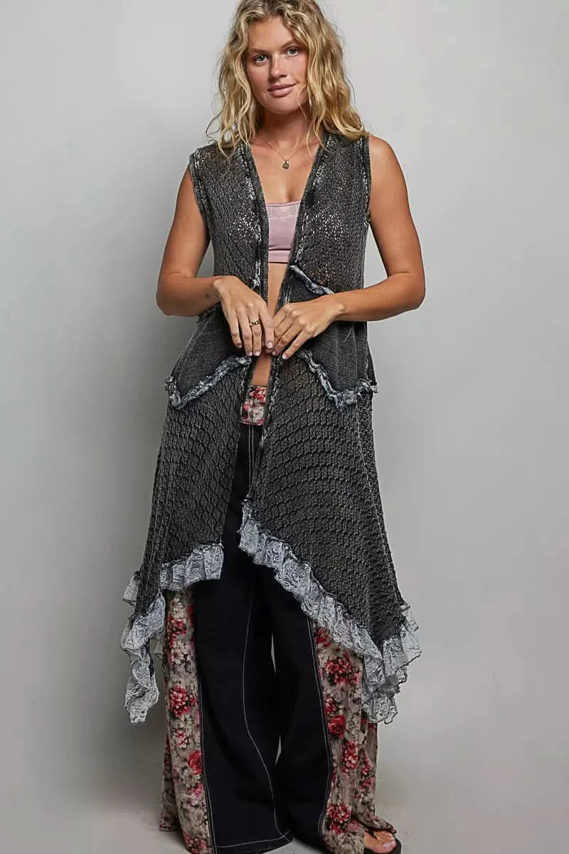 Woman wearing a vintage washed black sleeveless open crochet vest with mixed knit textures, lace ruffle trim, and an asymmetrical handkerchief hem layered over a neutral bralette and dark denim.