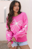 Model wearing a pink sweater with white embroidered bow design, standing against a light background