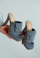 Pair of denim high-heeled shoes with pouch pocket held by a hand on a light gray background