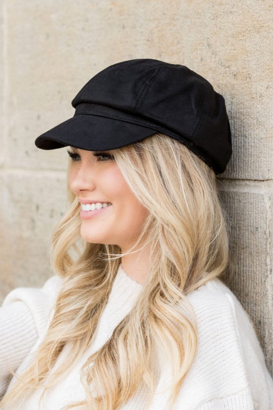 Black faux suede newsboy cap with structured bill worn by model smiling in profile
