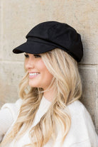 Black faux suede newsboy cap with structured bill worn by model smiling in profile
