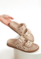 Neutral beige faux leather sandal with metallic studs and adjustable buckles displayed on white surface