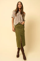 Model standing with hands in pockets wearing olive midi denim skirt and floral blouse.
